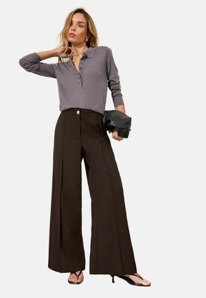 Femeie purtând un top gri cu mâneci lungi cu nasturi, pantaloni maro largi și cu talie înaltă, sandale negre și ținând o poșetuță neagră.
