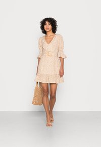 Vestido com padrão floral em bege, com decote em V, mangas bufantes e bainha rodada. Combinado com uma bolsa de palha e sapatos de salto agarre.