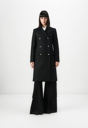 Woman with straight dark hair wears a black double-breasted coat with gold buttons and wide-legged black pants, standing against white background.