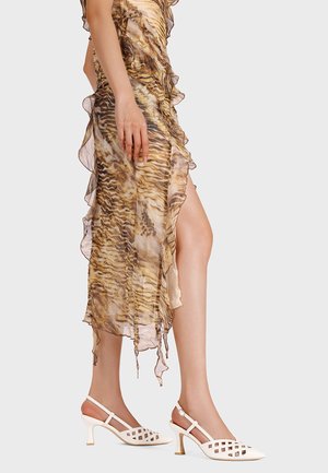 Donna che indossa un vestito trasparente con stampa tigre, volant e spacco alto sulla coscia, abbinato a sandali slingback bianchi con motivo a rete.