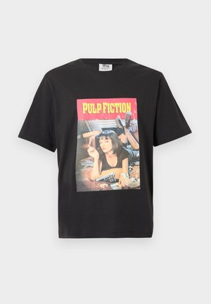 Melna T-kreklā ar grafisku attēlu, kurā sieviete guļ gultā, tur cigareti, blakus gultai atrodas pistole, un virs tā ir teksts "PULP FICTION".