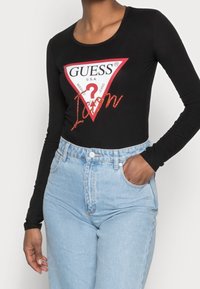 Czarna bluzka z długim rękawem z czerwono-białym trójkątnym logo z napisem "GUESS" i "Icon" w cekinach, zestawiona z jasnoniebieskimi spodniami o wysokim stanie.