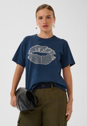 Femme portant un t-shirt bleu marine avec une empreinte de lèvres blanche, un pantalon vert olive, tenant une pochette en cuir noire et des boucles d'oreilles géométriques.