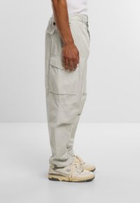 Pantaloni cargo grigio chiaro con multiple tasche, vestibilità slim e tessuto resistente. Indossati con sneakers beige con accenti e lacci grigi.