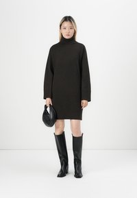 Tummanruskea neulemekko, jossa on korkea kaula-aukko, pitkät hihat ja ribbaushelma, yhdistetty mustiin nahkasaappaisiin polven ylle ja mustaan laukkuun.