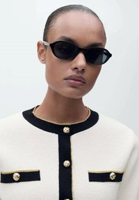 Des lunettes de soleil en forme de chat noir avec des accents argentés, portées avec un cardigan crème, noir et or avec des boutons dorés et un tissu texturé.