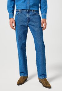 Mies wearing blue denim pants, matching denim shirt, and brown cowboy boots standing against a plain white background.