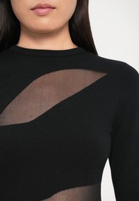 Top nero a maglia con inserti in rete trasparente a motivo ondulato, collo rotondo e maniche lunghe. Texture liscia con dettagli delicati.