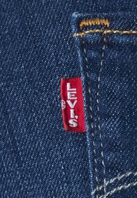 Tela de mezclilla azul oscuro con costuras en un amarillo dorado; presenta una etiqueta roja de Levi's con texto blanco cosida en la costura lateral.