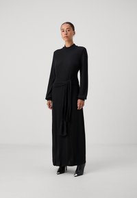 Črna dolga obleka s prožnimi teksturami, visokim ovratnikom in detajlom pasu s kravato; v kombinaciji z ostro obutvijo v črni barvi. Minimalistična zasnova.