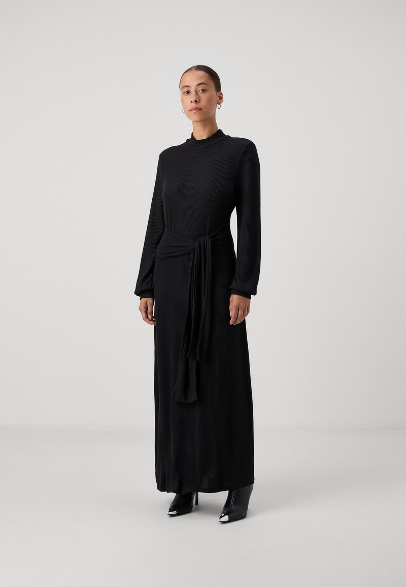 Črna dolga obleka s prožnimi teksturami, visokim ovratnikom in detajlom pasu s kravato; v kombinaciji z ostro obutvijo v črni barvi. Minimalistična zasnova.