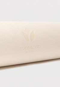 Gros plan sur un tapis de yoga beige texturé avec un logo en relief Even & Odd Active et un motif abstrait de feuille.