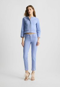 Gaiši zila īsa denim jaka ar priekšējām kabatām un ziedu rotājumiem, kombinēta ar atbilstošiem šauriem biksēm un bēšām siksniņu kurpēm.