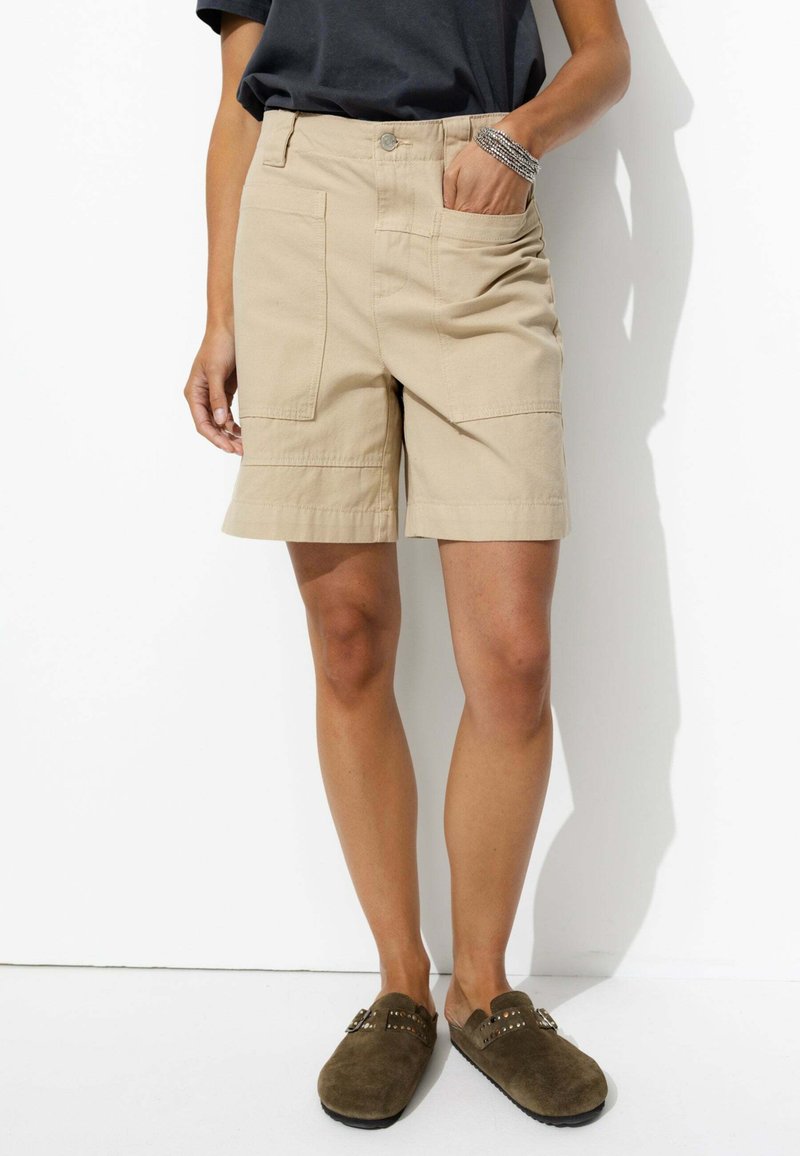 Bermuda cargo beige avec une coupe régulière, dotée de deux poches avant et d'un ourlet légèrement roulé. Associée à des sabots en suède brun foncé.