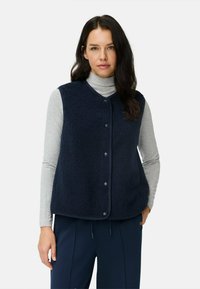 Marineblauwe fleece vest met een gestructureerd oppervlak, voorzien van een V-hals en drukknoopsluitingen, gedragen over een grijze coltrui. Losse pasvorm.