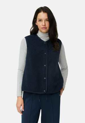 Marineblauwe fleece vest met een gestructureerd oppervlak, voorzien van een V-hals en drukknoopsluitingen, gedragen over een grijze coltrui. Losse pasvorm.