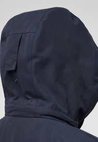 Marineblauwe waterdichte jas met capuchon en een gladde textuur, voorzien van een achterflap en stikdetails. Inclusief een drukknoop onder de flap.