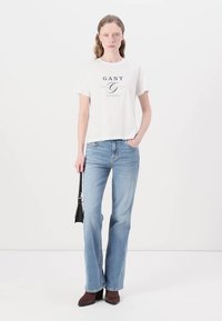 T-shirt bianco in cotone con stampa blu navy, abbinato a jeans flare in denim azzurro chiaro. Accentuato con una borsa nera e stivali con tacco marroni.