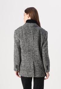 Blazer gris et noir à motif pied-de-poule avec un col haut, des épaules structurées et des poignets à deux boutons. Accompagné de pantalons noirs ajustés.