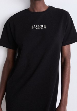 Osoba nosząca czarną koszulkę z "Barbour International" białym tekstem wyśrodkowanym na piersi na jednolitym tle.