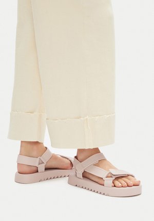 Lekko różowe sandały na platformie z podeszwą w zygzak, regulowanymi paskami i błyszczącą cholewką. Noszone z kremowymi szerokimi spodniami o postrzępionym dole.