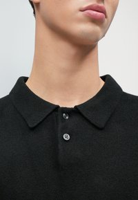 Polo shirt in lana nera con colletto piatto, dotato di bottoni e tessuto testurizzato. Vista ravvicinata che mette a fuoco il collo e i bottoni.