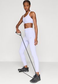 Ανοιχτό μωβ αθλητικό σουτιέν και αντίστοιχο κολάν με ψηλή μέση, και τα δύο από ελαστικό υλικό. Το μοντέλο κρατά μια μαύρη ζώνη αντίστασης με λαβές.