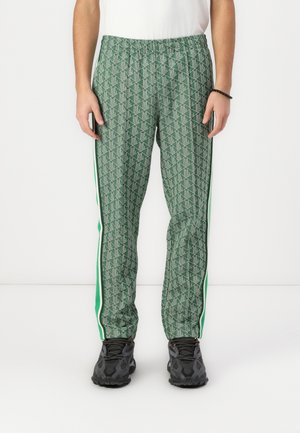 Personne portant un pantalon vert à motif géométrique avec des bandes latérales blanches et vertes, des baskets noires et un haut blanc sur un fond uni.