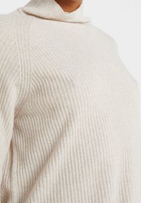 Close-up van een persoon die een lichtbeige ribbelgebreide coltrui draagt, waarbij textuur en stofdetails zichtbaar zijn.