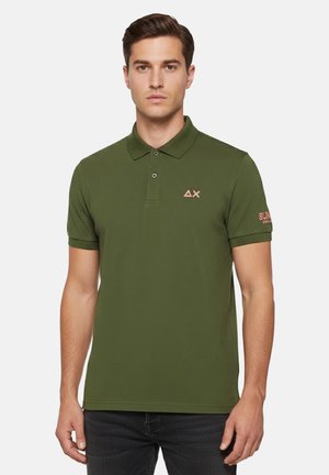 Uomo che indossa una polo verde oliva a maniche corte con piccolo logo sul petto e testo sulla manica sinistra, in piedi contro uno sfondo bianco semplice.