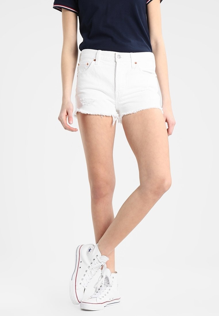 Witte denim shorts met rafelige zoom, hoge taille en subtiele distressed details, gecombineerd met witte hoge sneakers en een marineblauw shirt.