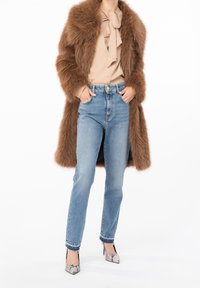 Abrigo de piel sintética marrón, blusa beige con detalle de lazo, jeans de mezclilla azul claro y tacones gris con estampado de serpiente. Enfoque en las texturas y los colores contrastantes.