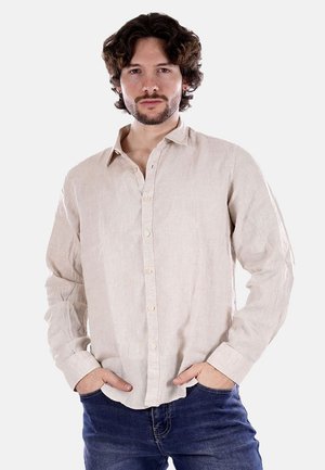 Uomo con capelli ricci che indossa una camicia beige con bottoni e jeans blu, in piedi con le mani nelle tasche contro uno sfondo uniforme.