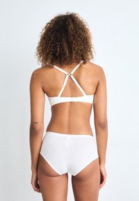 Witte katoenen bralette met kruislings straps en haaksluiting, gecombineerd met bijpassende high-waisted slips, gepresenteerd tegen een effen achtergrond.