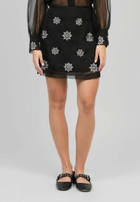 Jupe noire avec des motifs floraux brodés en argent et blanc. Longueur au-dessus du genou, faite d'un matériau léger, associée à des chaussures plates noires.