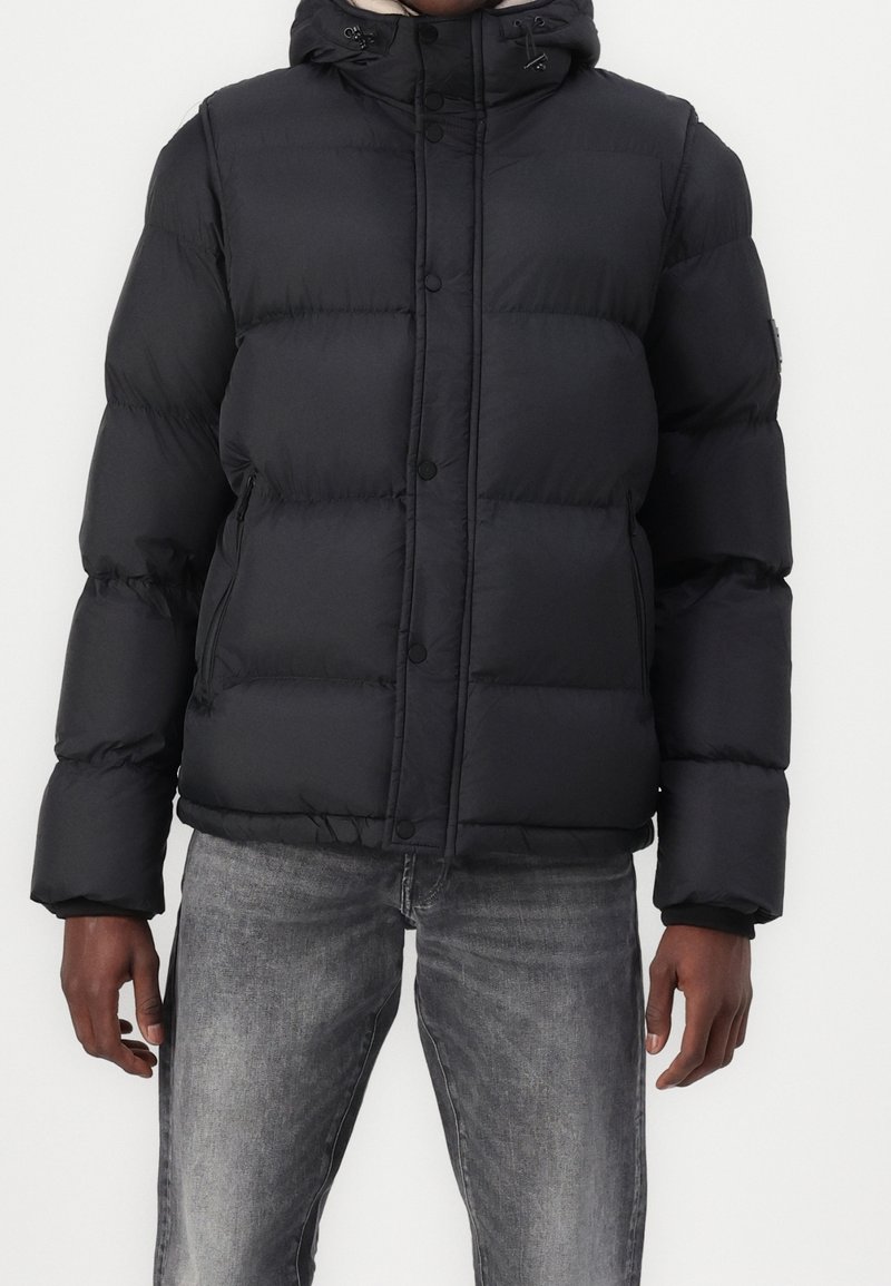 Homme portant une doudoune noire à capuche et un jean gris délavé, debout devant un fond blanc uni.