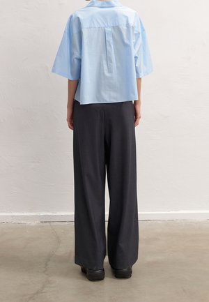 Person standing with back facing camera wearing a light blue short-sleeve shirt and loose black pants on a concrete floor.