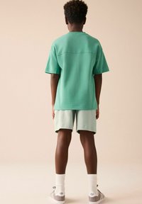 Grøn bomulds t-shirt med korte ærmer og rund halsudskæring, parret med lysegrønne shorts. Tilbehøret omfatter hvide sokker og grå sneakers.