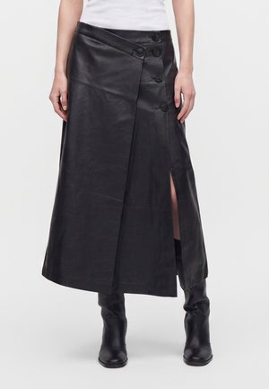 Jupe midi en cuir noir avec détails de boutons et fente à l'avant, portée avec des bottes noires et un top blanc.