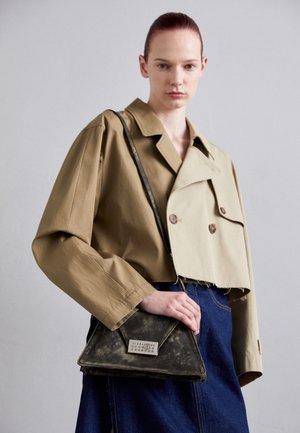 Mujer con cabello rojo lleva una chaqueta corta bicolor beige, falda azul oscura y sostiene un bolso triangular negro desgastado con un cierre plateado.