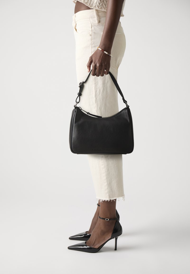 Black leather handbag with a curved design, adjustable strap, and zip closure. Model wears cream pants and black pointed-toe heels.
