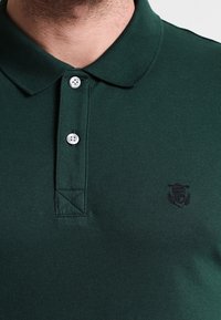 Ciemnozielona koszulka polo z teksturowanej tkaniny, z trzema guzikami przy kołnierzyku oraz małym czarnym haftem logo po lewej stronie.