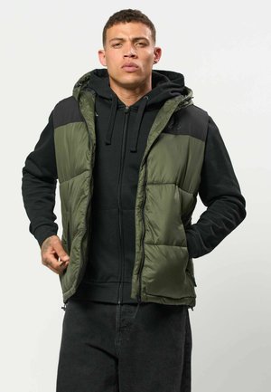 Junger Mann trägt schwarzen Hoodie und grüne gesteppte Weste, stellt den Reißverschluss der Weste ein, steht vor schlichtem hellem Hintergrund.