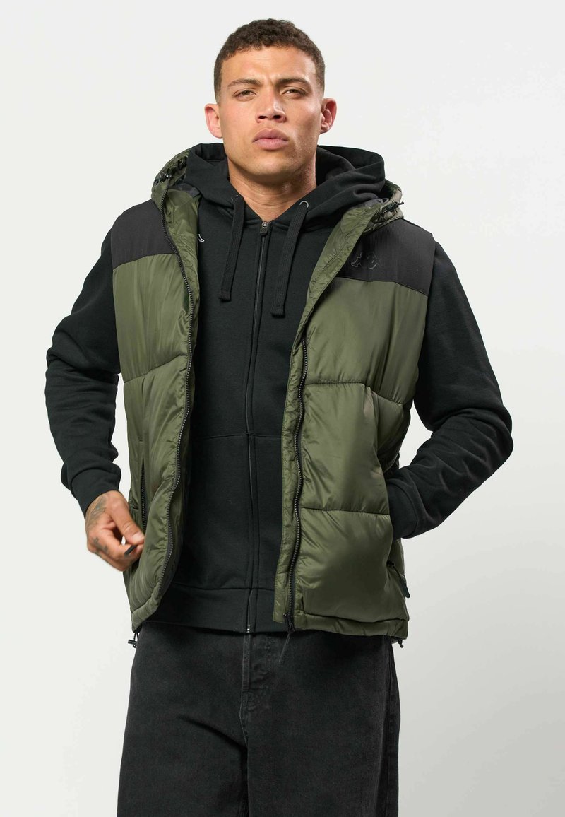 Jeune homme portant un sweat à capuche noir et un gilet matelassé vert, ajustant la fermeture éclair du gilet, debout devant un fond clair uni.