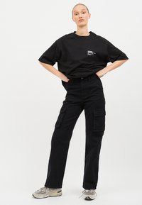 Schwarzes Baumwoll-T-Shirt in Übergröße mit Brustdruck, kombiniert mit schwarzen Cargo-Hosen mit seitlichen Taschen und geradem Beinschnitt.