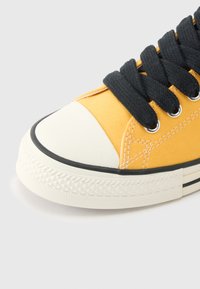 Κοντινή λήψη κίτρινου πάνινου αθλητικού παπουτσιού με μαύρα κορδόνια, λευκό καουτσούκ πλώρης και υφή λευκής σόλας σε ανοιχτό φόντο.