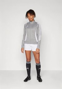 Γκρι αθλητικό μπουφάν με λευκές λεπτομέρειες, μπροστινό φερμουάρ και κλασικές τρεις ρίγες. Συνδυάζεται με λευκά σορτς και μαύρες ψηλές κάλτσες, που φέρουν το λογότυπο της μάρκας.