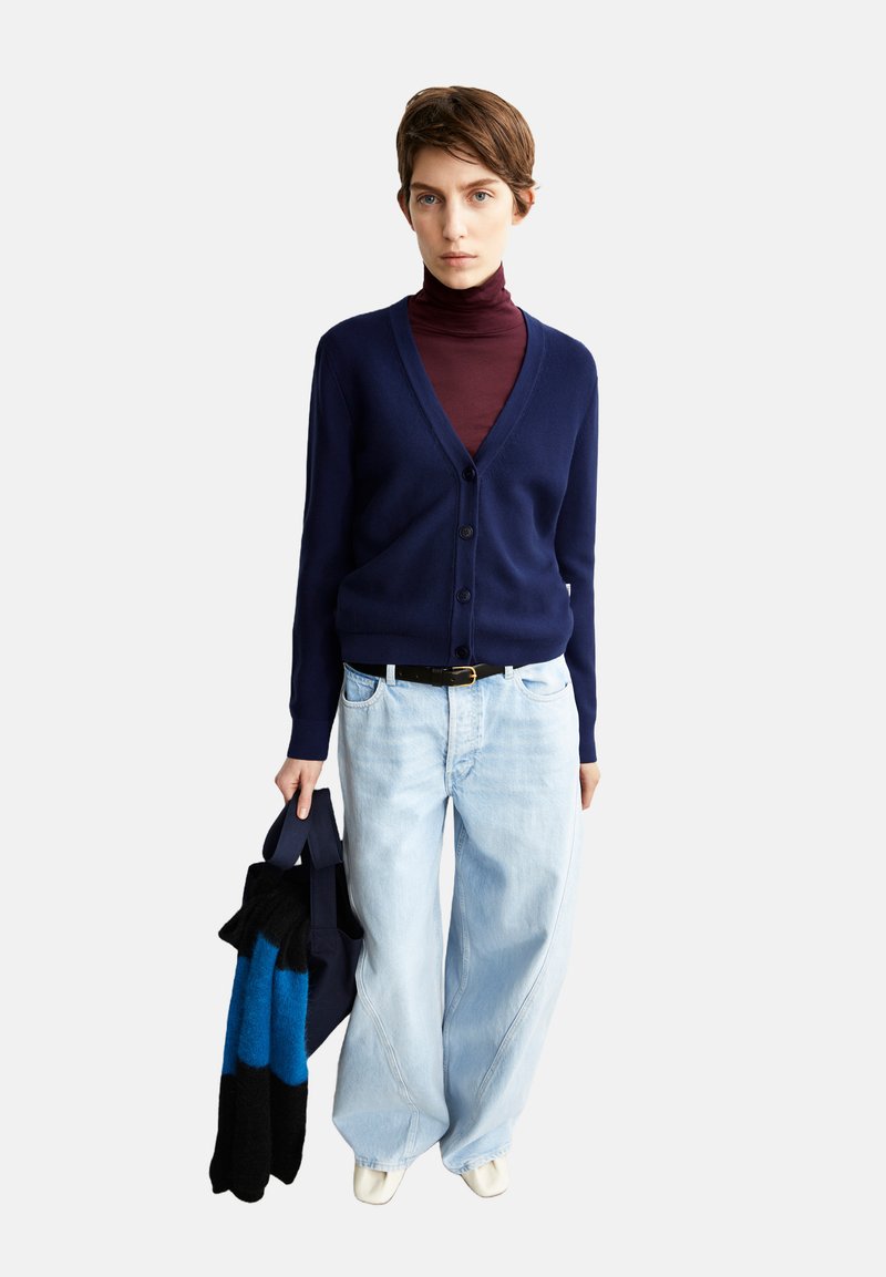 Marinefarbener Cardigan mit V-Ausschnitt und Knöpfen, getragen über einem weinroten Rollkragenpullover. Hellblaue, weit geschnittene Jeans und weiße Schuhe. Hält eine Tragetasche und einen Schal.