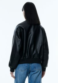 Schwarze Lederbomberjacke mit gerippten Bündchen und Saum, die eine glatte, glänzende Textur aufweist, kombiniert mit blauen Jeans.