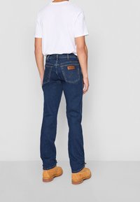 Vaqueros de mezclilla azul oscuro con corte recto, que presentan dos bolsillos traseros con una etiqueta de cuero marrón y detalles de costuras visibles. Combinados con botas de color cuero tostado.